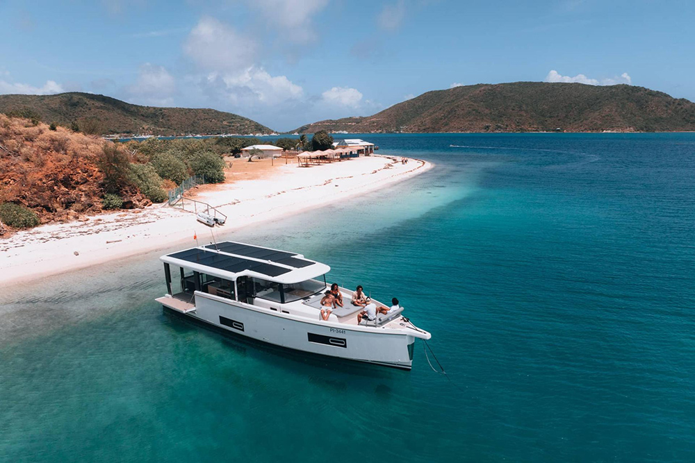 Jeanneau Sea Loft cruising beside a tropical beach, with solar panels on the roof and passengers relaxing at the bow.