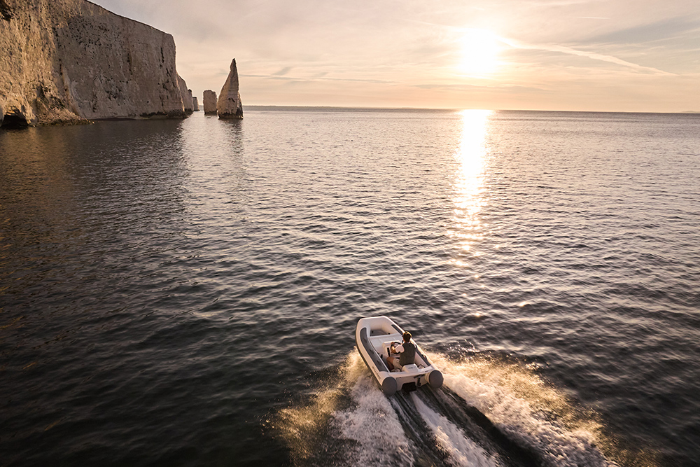 Jet tender speeding past chalk cliffs and sea stacks at sunset, golden light reflecting on calm water.