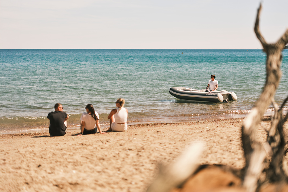 Williams jet tender approaching a sandy beach as three people watch from the shore, turquoise sea beyond.