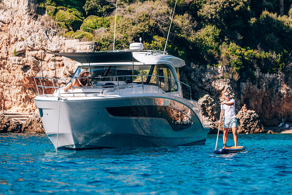 Beneteau Antares 12 motorcruiser at an chore with paddleboarder to the side and tree'd cliffs in the background