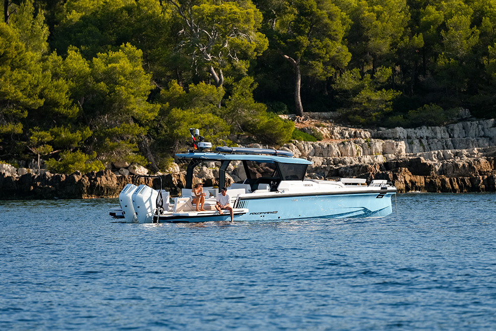 Axopar 38 Cross Top with twin outboards cruising a wooded coastline, two people relaxing on the open deck.