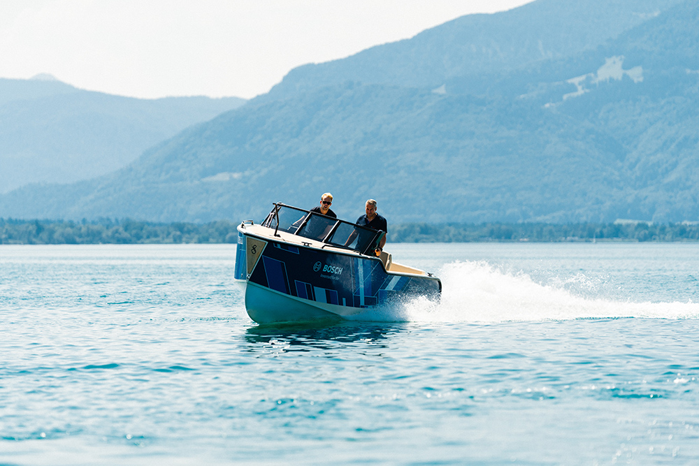 Bosch-branded electric boat planes across a lake, throwing spray, with two people aboard and mountains behind.