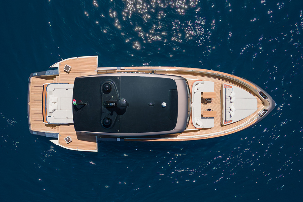 Aerial view of a motor yacht with teak decking, black hardtop and cushioned sun pads cruising sunlit blue water.