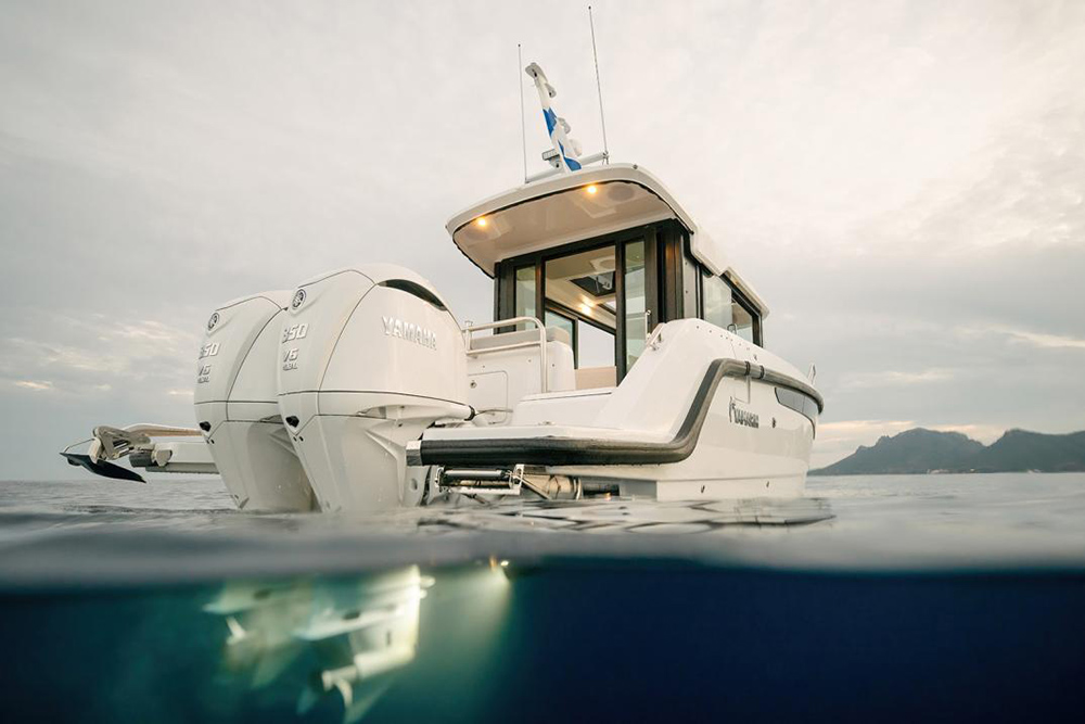 Yamarin Aura Cabin pilothouse with twin Yamaha F350 outboards, viewed at water level against a mountainous coastline.