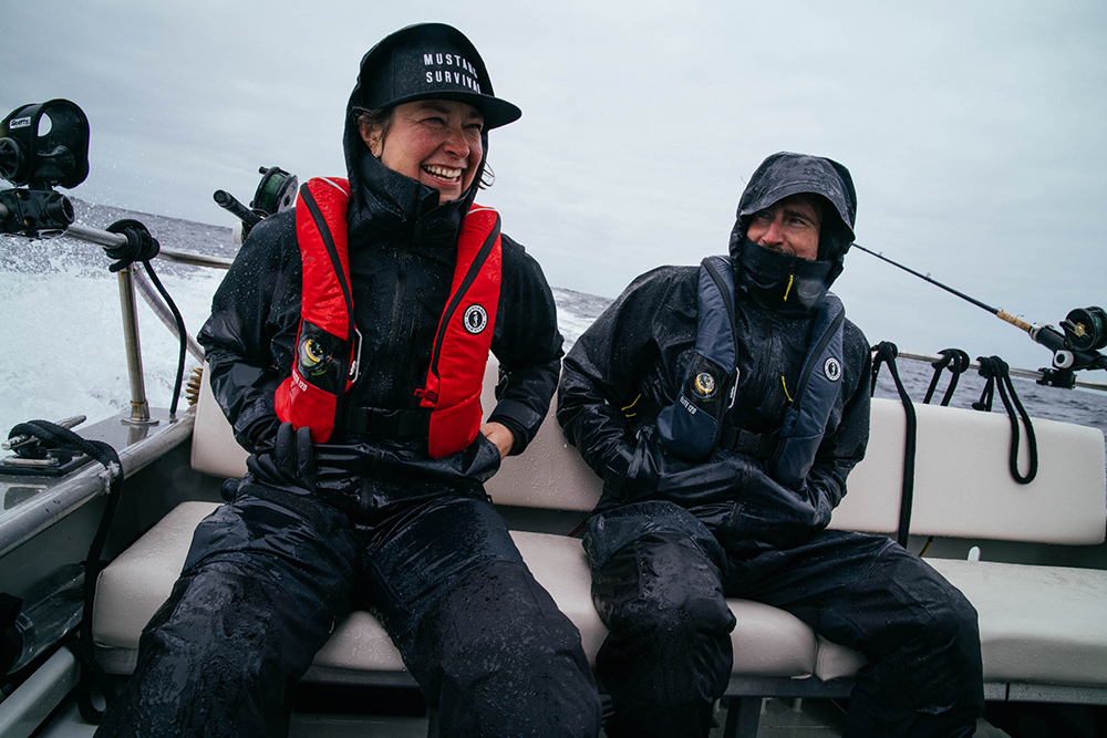 Two anglers wearing Mustang Survival Taku jackets and life vests on a boat, spray and a rod visible behind them.