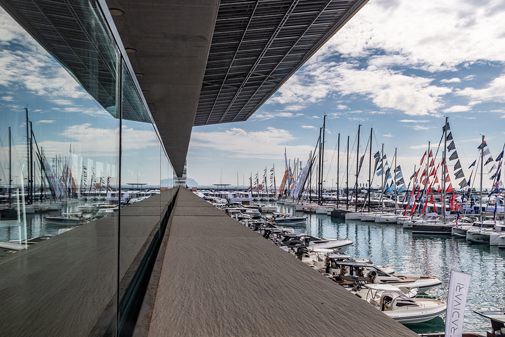 RIBs and sailing yachts moored at the Genoa International Boat Show marina, reflected in the glass of a waterfront pavilion