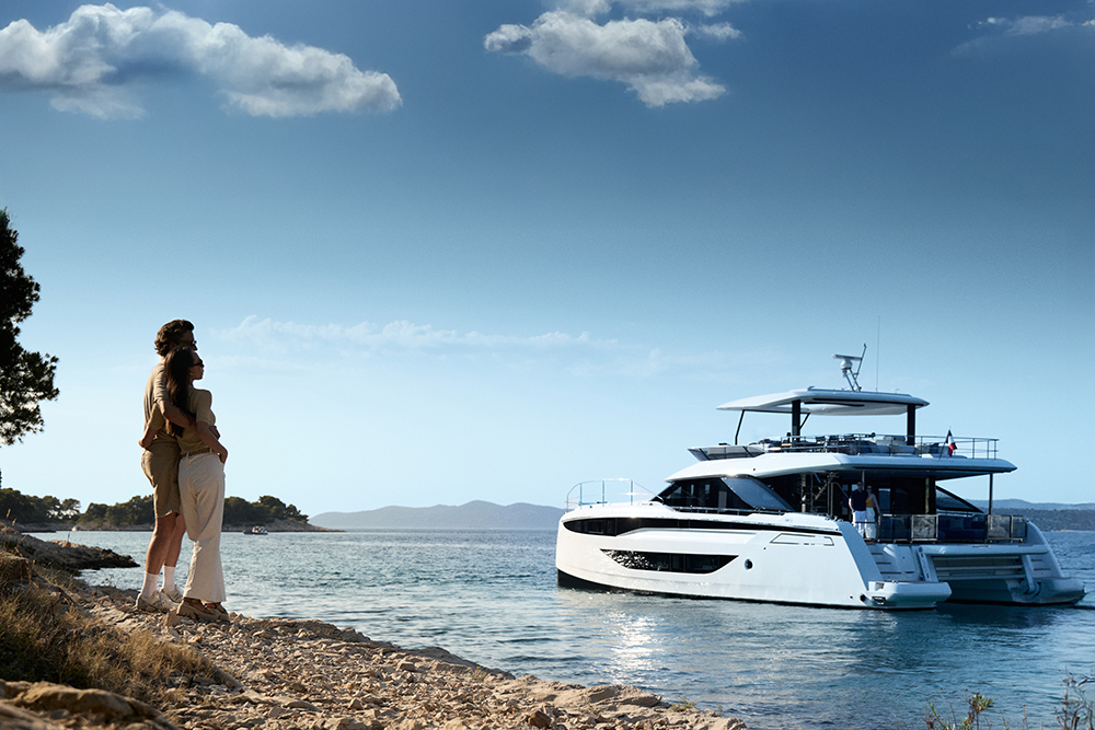Couple embracing on a rocky shoreline as the Prestige M8 EVO sits in a calm, sunlit bay.