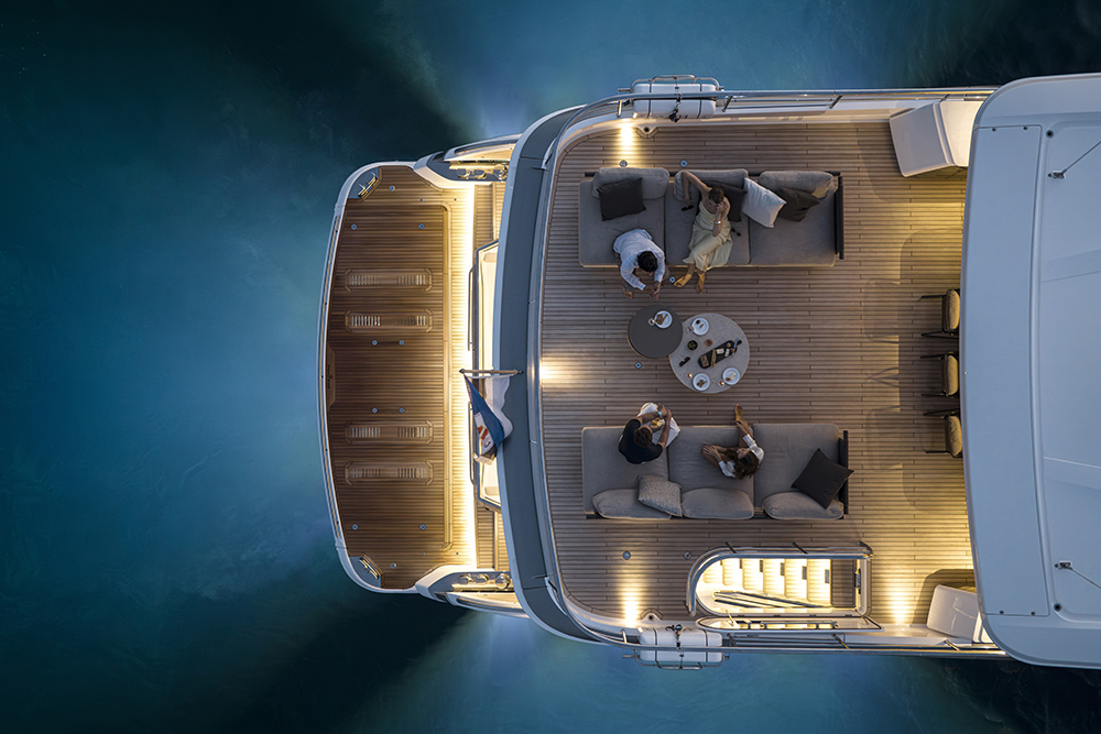 Aerial view of the Princess Yachts X90, a luxury motor yacht’s teak aft deck at dusk, with guests relaxing on sofas beside a lit swim platform