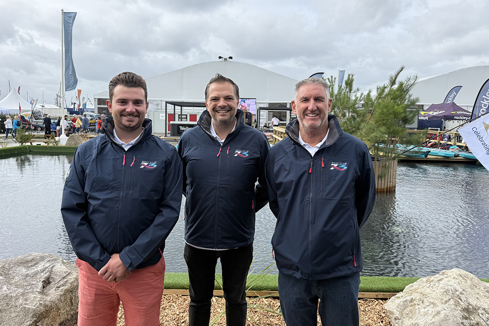 Three Boat Club 73 team members in branded navy jackets stand beside the water at a boat show, with marquees behind