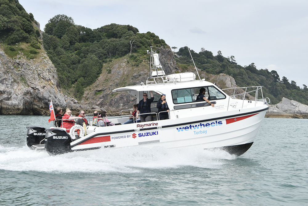 Wetwheels Torbay accessible powerboat with twin Suzuki outboards carrying passengers past wooded cliffs on the Devon coast.