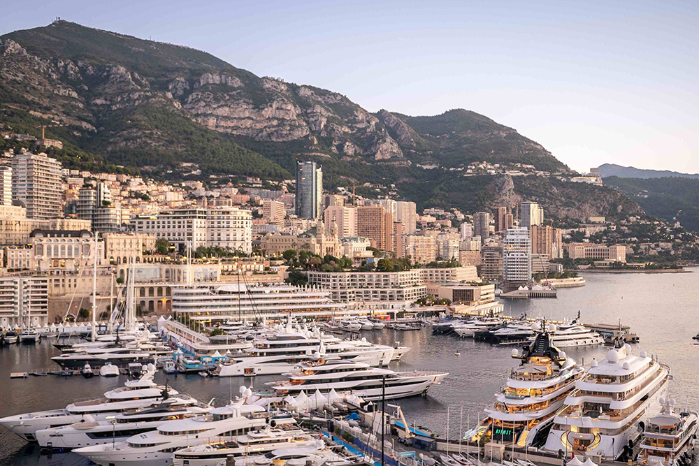Superyachts fill Port Hercule at dusk, with Monaco's hillside skyline and mountains rising behind the marina.